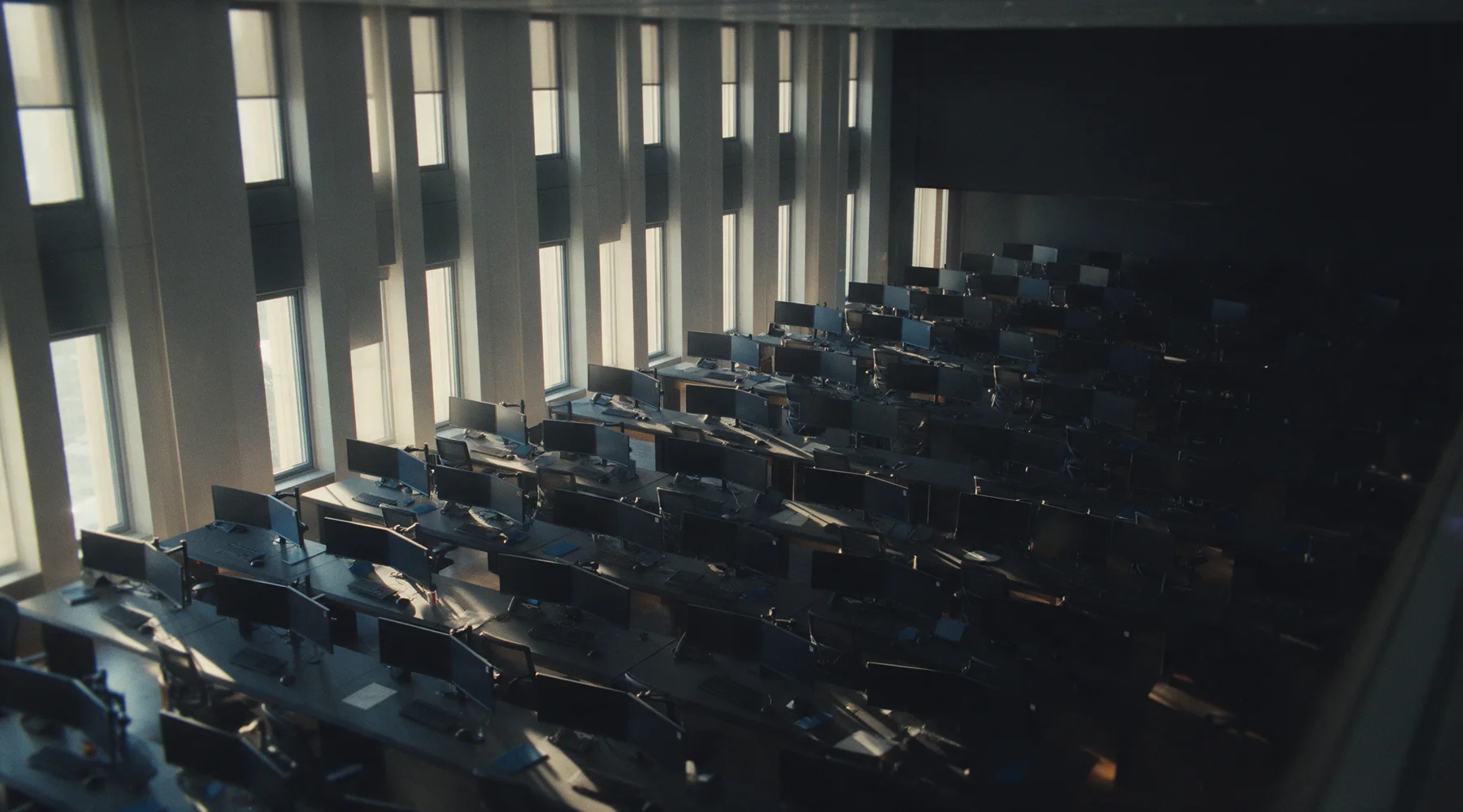 Rows of empty desks and dark monitors on a trading floor, low sun through tall windows.