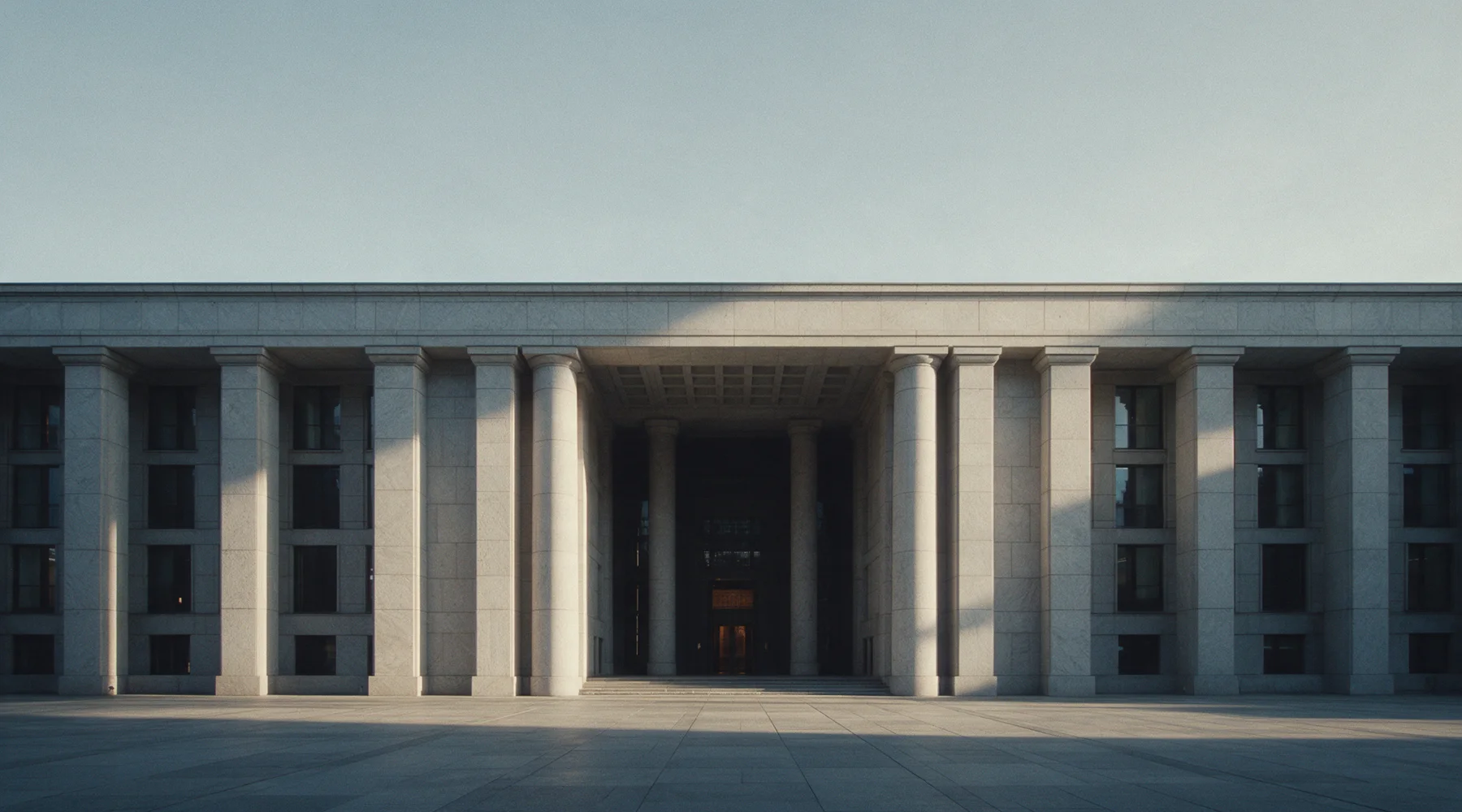 A long colonnaded stone facade above an empty paved forecourt, early light across the columns.